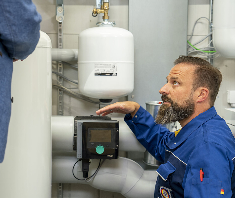 Eine erfahrene SHK-Fachkraft erklärt die Komponenten einer zentralen Heizungs- oder Warmwasserbereitungsanlage. Neben ihm steht ein Bauherr, Planer oder Energieberater.