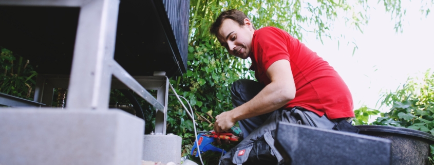Ein männlich gelesener SHK-Anlagenmechaniker baut in einem Garten eine Außeneinheit einer Wärmepumpe ein.