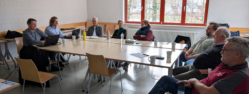Acht Personen sitzen in einem Workshop-Raum um Tische und arbeiten zusammen.